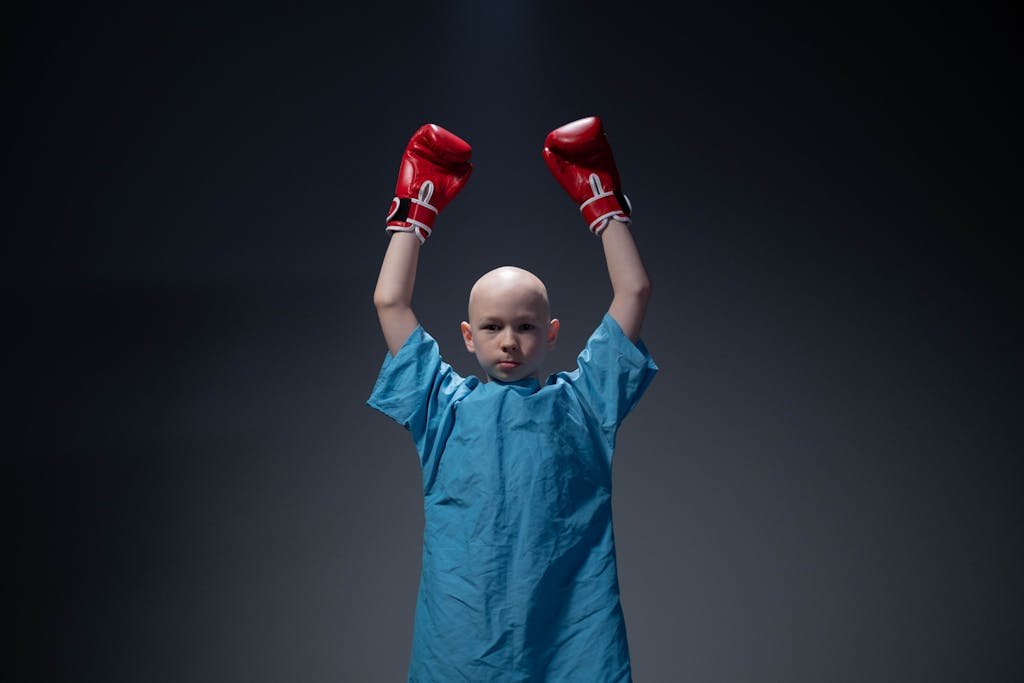 A child in a hospital gown and boxing gloves raises arms in victory, symbolizing strength and resilience.