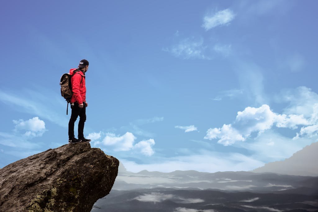 Hiker in red jacket stands on a cliff edge, overlooking a scenic mountain view under a bright blue sky.