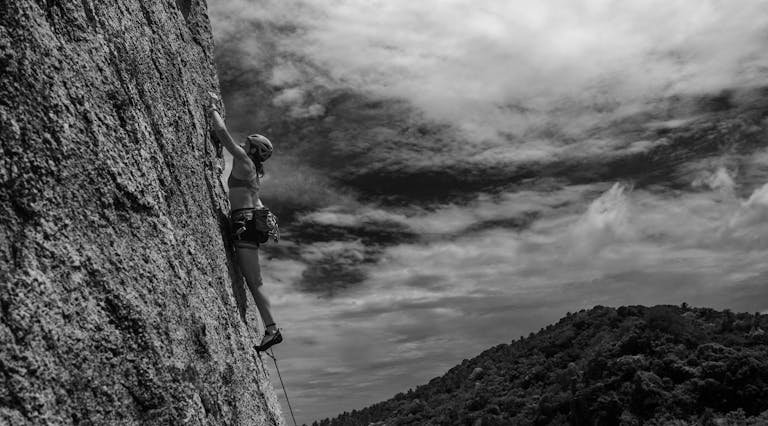 Woman climber scaling a steep rock face under an overcast sky, embodying adventure and determination.